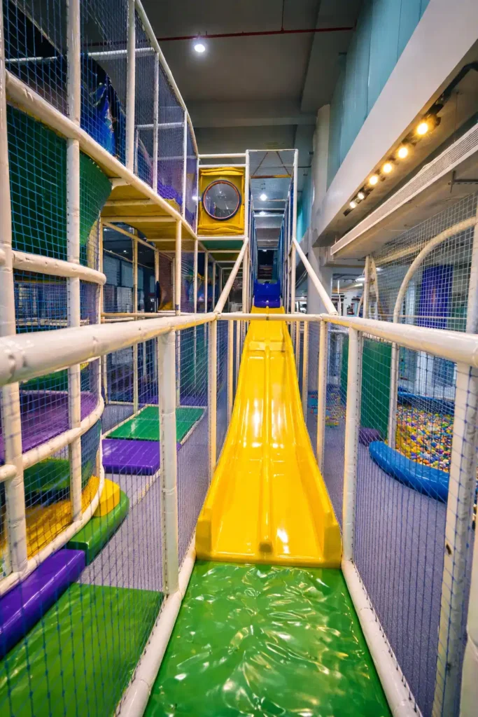 ممر ألعاب داخلي يقود إلى زحليقة صفراء داخل مركز ترفيهي للأطفال يحتوي على ألعاب تسلق ومسبح كرات.