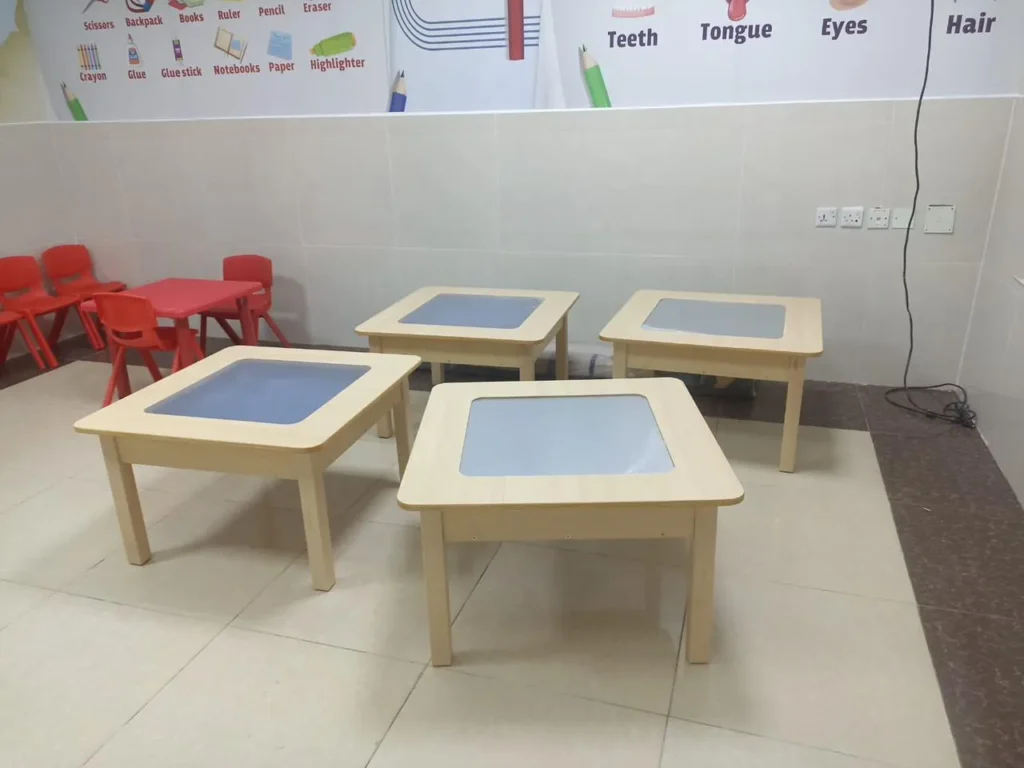 طاولات تعليمية خشبية مربعة للأطفال بألوان فاتحة ومحيط تعليمي مزين برسومات أدوات الدراسة