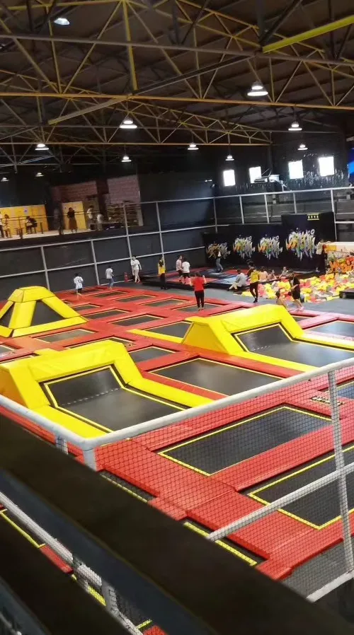صالة العاب داخلية تحتوي على مجموعة من الترامبولين الملونة بالأصفر والأحمر، حيث يلعب الأطفال تحت سقف مضاء، مع وجود منطقة إسفنجية للأمان