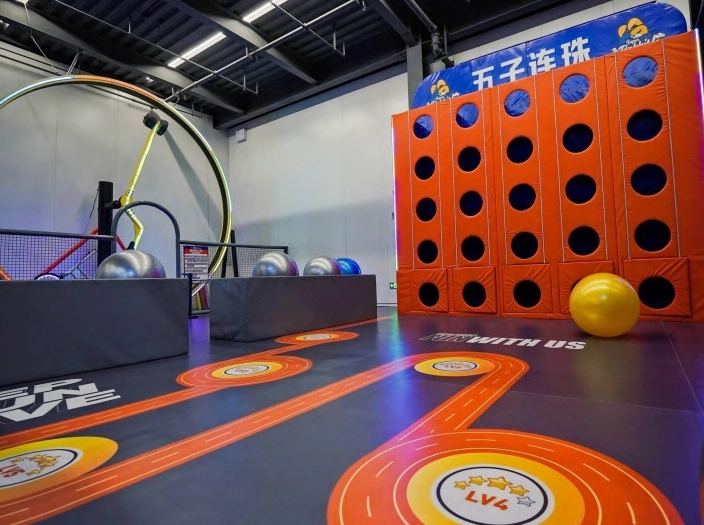 منطقة ألعاب حركية داخل صالة ترفيهية تضم جدار تحدي برتقالي بفتحات تسلق وأرضية تفاعلية ملونة مع مسارات مرسومة وكرات كبيرة