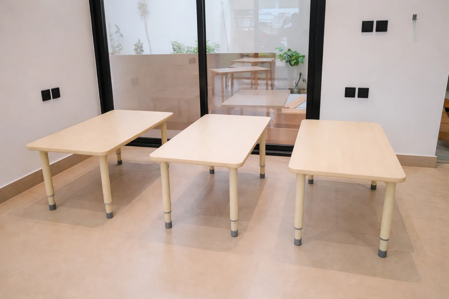 طاولات دراسية مستطيلة للأطفال بتصميم بسيط داخل فصل حديث مع أرضية نظيفة وواجهة زجاجية تطل على ساحة خارجية.